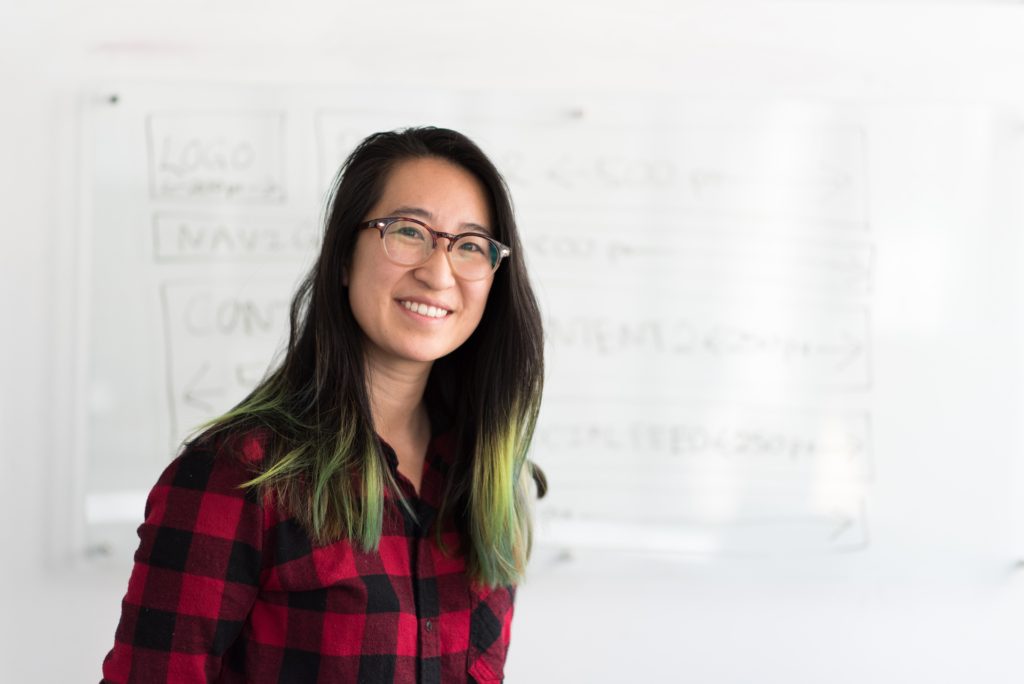 pretty teacher with a white board behind her. teacher has glasses on and a black and red plaid shirt. jo & co realty group program for teachers only in klein isd and all of houston